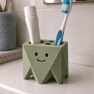 May include: A sage green geometric toothbrush holder with a smiling face design, featuring multiple circular openings for storage. A white toothpaste tube and a white and blue toothbrush are inserted into the top of the holder.