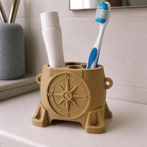 Pode incluir: Um suporte para escovas de dentes bege em forma de barril, com um design de rosa dos ventos em relevo na frente, exibindo os pontos cardeais N, S, E e O. O suporte possui quatro aberturas circulares, contendo um tubo branco e uma escova.
