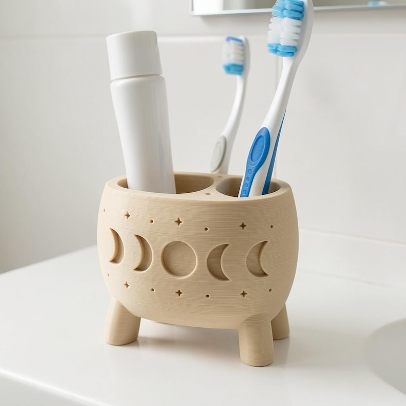 May include: A beige, three-legged toothbrush holder featuring a cutout moon phase pattern and small star motifs. The holder contains a white tube and two toothbrushes with blue and white bristles, providing storage for bathroom essentials.