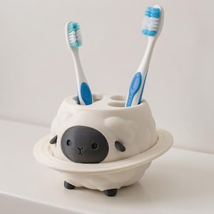 May include: A white toothbrush holder shaped like a sheep with a dark gray face, small ears, and four small feet. The holder features a circular ring around the middle and has four holes on top, two of which contain blue and white toothbrushes.