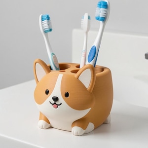 Puede incluir: Un soporte para cepillos de dientes con forma de Corgi en color tostado y blanco, con tres aberturas circulares en la parte superior. El soporte presenta una cara de perro de dibujos animados con ojos negros, nariz negra y una peque&ntilde;a lengua rosa.