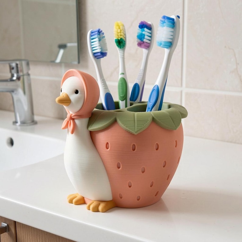 May include: A white duck figurine wearing a pink bonnet attached to a peach-colored strawberry-shaped toothbrush holder with green leaves at the top. The holder contains four toothbrushes with blue, yellow, purple, and white bristles.