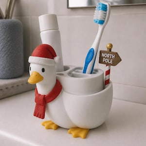 May include: A white duck-shaped toothbrush holder featuring a red Santa hat, a red scarf, and yellow feet. The holder includes a small signpost labeled NORTH POLE, a white tube of toothpaste, and a blue and white toothbrush in the storage compartments.