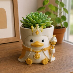 May include: A white and yellow planter shaped like a duck wearing a crown and a cape. The duck holds a small scepter topped with a duck head and wears a medallion featuring a duck face. A green succulent plant is potted inside the duck-shaped container.