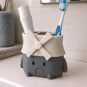 May include: A gray and off-white toothbrush holder shaped like a windmill with a brick-textured base. The holder features a central windmill blade design on the front and multiple compartments containing a white toothpaste tube and a blue toothbrush.
