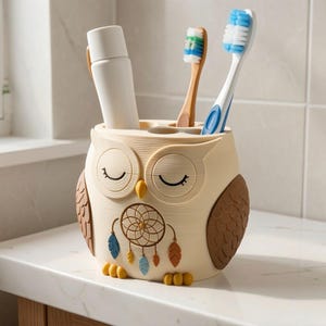 May include: A cream-colored owl-shaped toothbrush holder with brown wings and yellow feet. The owl features closed eyes, a yellow beak, and a dreamcatcher graphic on its chest with blue, yellow, and orange feathers. It holds a white tube and two toothbrushes.
