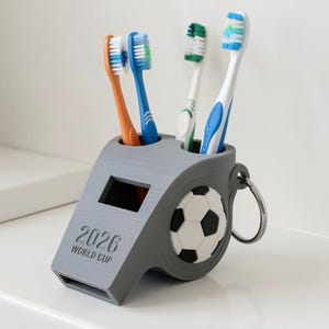 May include: A gray plastic toothbrush holder shaped like a sports whistle with a soccer ball emblem on the side. The holder features the text 2026 WORLD CUP and contains four toothbrushes with orange, blue, green, and blue-and-white bristles.