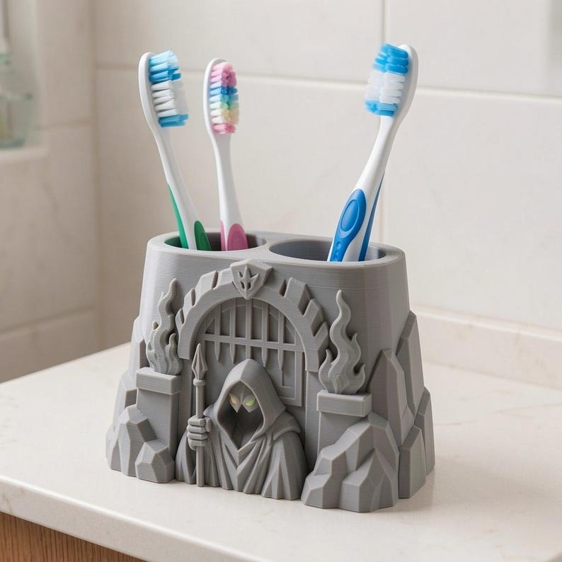 Puede incluir: Un soporte para cepillos de dientes de pl&aacute;stico gris con forma de puerta de fortaleza de piedra con una figura encapuchada que sostiene una lanza en el centro. El soporte tiene dos aberturas circulares en la parte superior con tres cepillos.