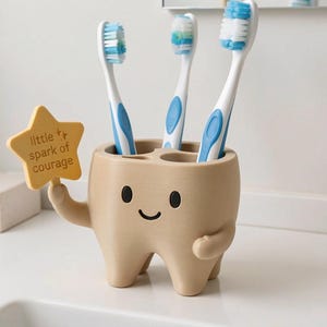 May include: A beige tooth-shaped toothbrush holder with a smiling face, holding a yellow star-shaped sign that reads little spark of courage. The holder contains three white and blue toothbrushes with blue bristles, standing upright in the slots.