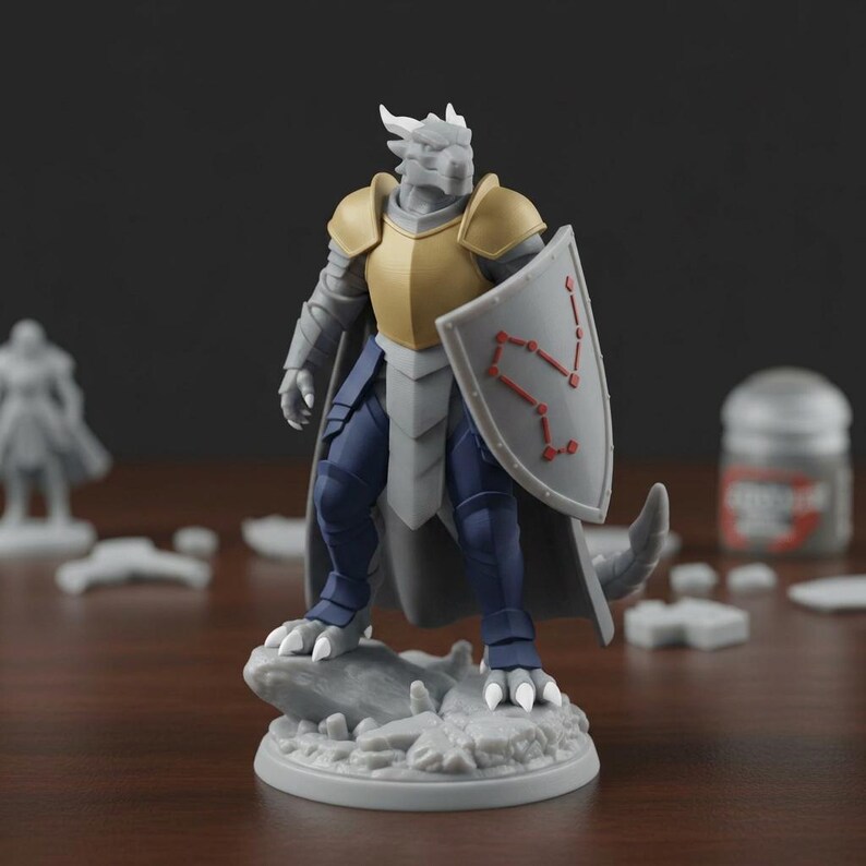May include: A gray and gold fantasy miniature figure of a dragon-like warrior, holding a shield with a red constellation design. The figure is posed on a rocky base, with blue pants and a gray cape. Other miniature figures and paint are in the background.