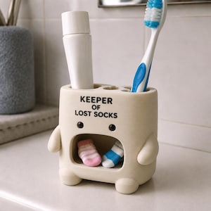 May include: A cream-colored, character-shaped toothbrush holder with the text KEEPER OF LOST SOCKS printed in black. The holder features a hollow center containing two miniature socks, with a white toothpaste tube and a blue toothbrush inserted.