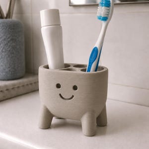 Pu&ograve; includere: Un portaspazzolino grigio chiaro con un design a faccina sorridente. Il supporto ha quattro scomparti ed &egrave; sostenuto da tre piccole gambe. Un tubetto di dentifricio bianco e uno spazzolino con dettagli blu sono all'interno.