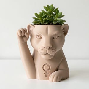 May include: A light beige planter shaped like a lioness with a raised paw in a fist gesture. The chest features a female gender symbol embossed on the surface. A green succulent plant is potted inside the top opening of the lioness figure.