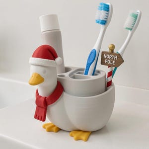 May include: A light gray duck-shaped toothbrush holder featuring a red Santa hat, a red scarf, and yellow feet. The holder contains a tube of toothpaste, two toothbrushes, and a small signpost with the text NORTH POLE in white lettering.