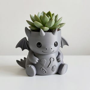May include: A matte gray 3D-printed dragon planter featuring a succulent plant. The dragon has large black eyes, small wings, and embossed icons on its belly including a diamond, a key, a paperclip, and a twenty-sided die. The surface is smooth.