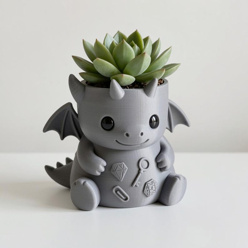 May include: A matte gray 3D-printed dragon planter featuring a succulent plant. The dragon has large black eyes, small wings, and embossed icons on its belly including a diamond, a key, a paperclip, and a twenty-sided die. The surface is smooth.