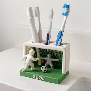 May include: A white and green plastic toothbrush holder shaped like a football goal with a net, featuring a white player figure, a grey goalkeeper figure, a small football, and the year 2026 printed on the front base, holding three toothbrushes.