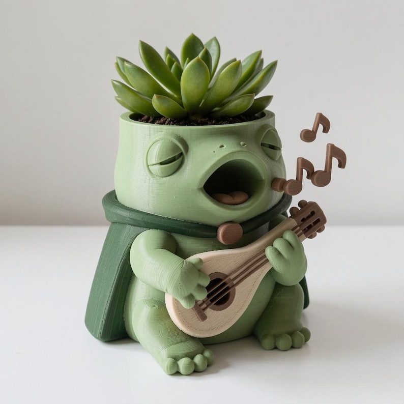 Puede incluir: Una maceta con forma de rana de color verde claro que contiene una planta suculenta. La rana tiene los ojos cerrados, la boca abierta y lleva una capa verde oscuro. Sostiene un la&uacute;d marr&oacute;n claro con tres notas musicales marrones al lado.