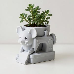 Peut inclure: Un pot de fleurs gris clair en forme de souris assise devant une machine &agrave; coudre. La souris a de grandes oreilles, des yeux noirs et des moustaches. Une plante succulente verte est plant&eacute;e dans la partie sup&eacute;rieure de la base.