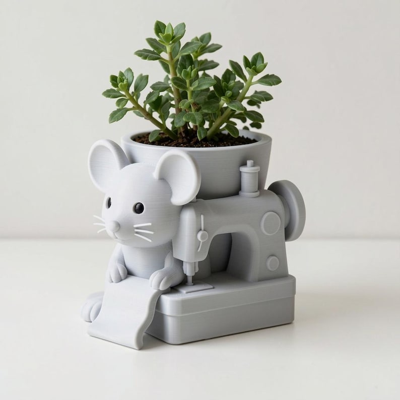 Peut inclure: Un pot de fleurs gris clair en forme de souris assise devant une machine &agrave; coudre. La souris a de grandes oreilles, des yeux noirs et des moustaches. Une plante succulente verte est plant&eacute;e dans la partie sup&eacute;rieure de la base.