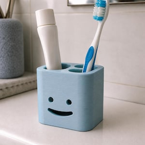 Puede incluir: Un porta cepillos de dientes azul claro con un dise&ntilde;o de cara sonriente. Contiene un tubo de pasta de dientes blanco y un cepillo de dientes blanco y azul. El soporte es de material texturizado y est&aacute; sobre una superficie blanca.