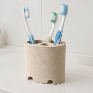 May include: A beige toothbrush holder with four slots, holding three toothbrushes. The toothbrushes have white handles with blue and green accents and colored bristles. The holder has a ribbed texture and a rounded rectangular shape.