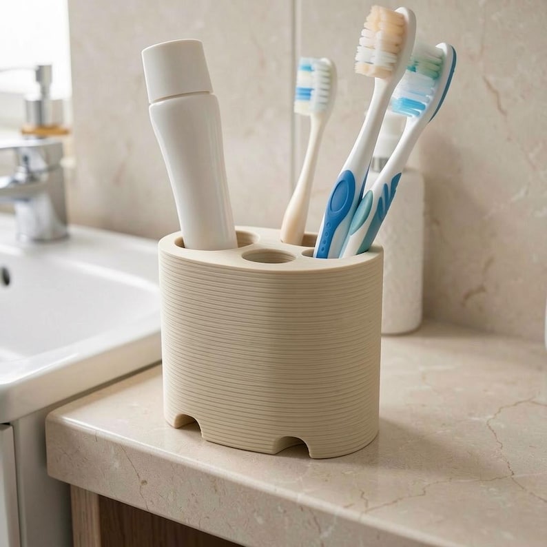 May include: A beige toothbrush holder with a tube of toothpaste and three toothbrushes. The toothbrushes have white handles with blue and green bristles. The holder is cylindrical with a ridged texture and four circular openings.