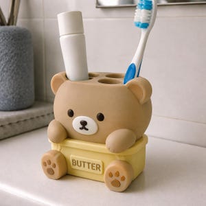 May include: A light brown bear-shaped toothbrush holder with a yellow base labeled BUTTER. The holder features a cartoon bear face, two small ears, and two front paws resting on the yellow base. It contains a white tube and a blue and white toothbrush.