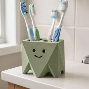 May include: A sage green, geometric-shaped toothbrush holder with a smiling face cutout. The holder features four circular openings on the top, currently containing four different toothbrushes with blue, green, white, and light blue bristles.