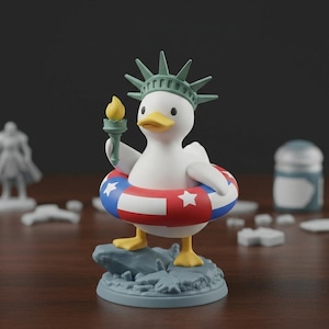 May include: A white duck figurine wearing a Statue of Liberty crown and holding a torch. The duck is wearing a red, white, and blue inflatable ring. The figurine is standing on a blue base with a rock-like texture.