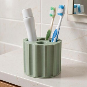 May include: A sage green toothbrush holder with a ribbed design. The holder contains a tube of toothpaste, and two toothbrushes with green and blue accents. The holder sits on a white countertop in a bathroom setting.