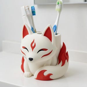 May include: A white fox-shaped toothbrush holder with red accents on the ears, forehead, and tail. The fox has closed eyes and a black nose. Two toothbrushes are inserted into the top openings of the holder, which is designed to store items.