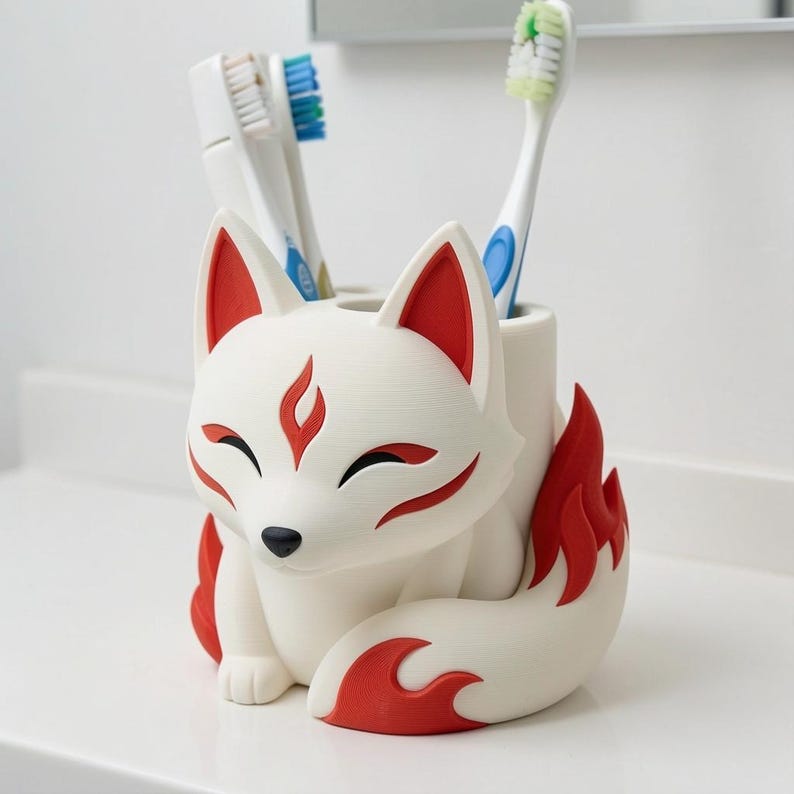 May include: A white fox-shaped toothbrush holder with red accents on the ears, forehead, and tail. The fox has closed eyes and a black nose. Two toothbrushes are inserted into the top openings of the holder, which is designed to store items.