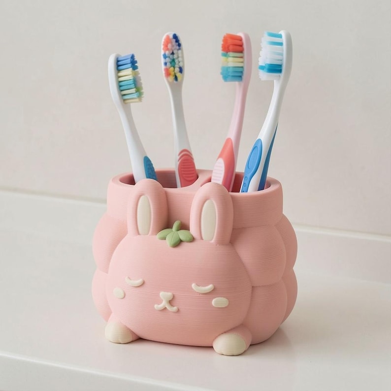 May include: A light pink toothbrush holder shaped like a rabbit with a small green leaf on top. The holder features a sleeping face with closed eyes and a small mouth. Four toothbrushes with white handles and multi-colored bristles are inserted.