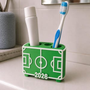 May include: A green and white toothbrush holder shaped like a soccer field, holding a white tube of toothpaste and a toothbrush with blue and white bristles. The number "2026" is printed on the front.