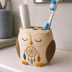 May include: A cream-colored owl-shaped toothbrush holder with brown wings, a yellow beak, and yellow feet. The front features a dreamcatcher graphic with blue, yellow, and orange feathers. It holds a white tube and a blue and white toothbrush.