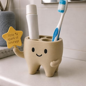May include: A beige tooth-shaped toothbrush holder with a smiling face, holding a yellow star-shaped sign that reads little spark of courage. The holder has four circular openings containing a white tube and a blue and white toothbrush.