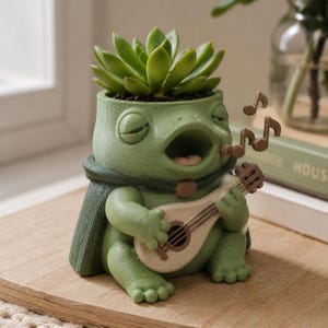 以下が含まれることがあります： 上部に多肉植物が植えられた、明るい緑色のカエルの形をしたプランター。カエルは目を閉じ、口を開けており、濃い緑色のマントを羽織って、小さな明るい色の弦楽器を演奏しています。周囲には音符の装飾が施されています。