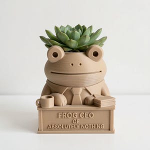 K&ouml;nnte beinhalten: Ein hellbrauner 3D-gedruckter &Uuml;bertopf in Form eines Frosches mit Krawatte, der an einem Schreibtisch sitzt. Auf dem Schreibtisch steht der Text "FROG CEO OF ABSOLUTELY NOTHING" sowie eine kleine Tasse und Papierstapel. Eine Sukkulente steckt im Kopf.