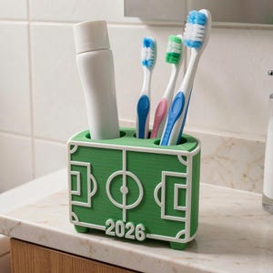 May include: A green and white toothbrush holder shaped like a soccer field, with the year "2026" printed on the front. The holder contains a tube of toothpaste and three toothbrushes with blue, pink, and green accents.