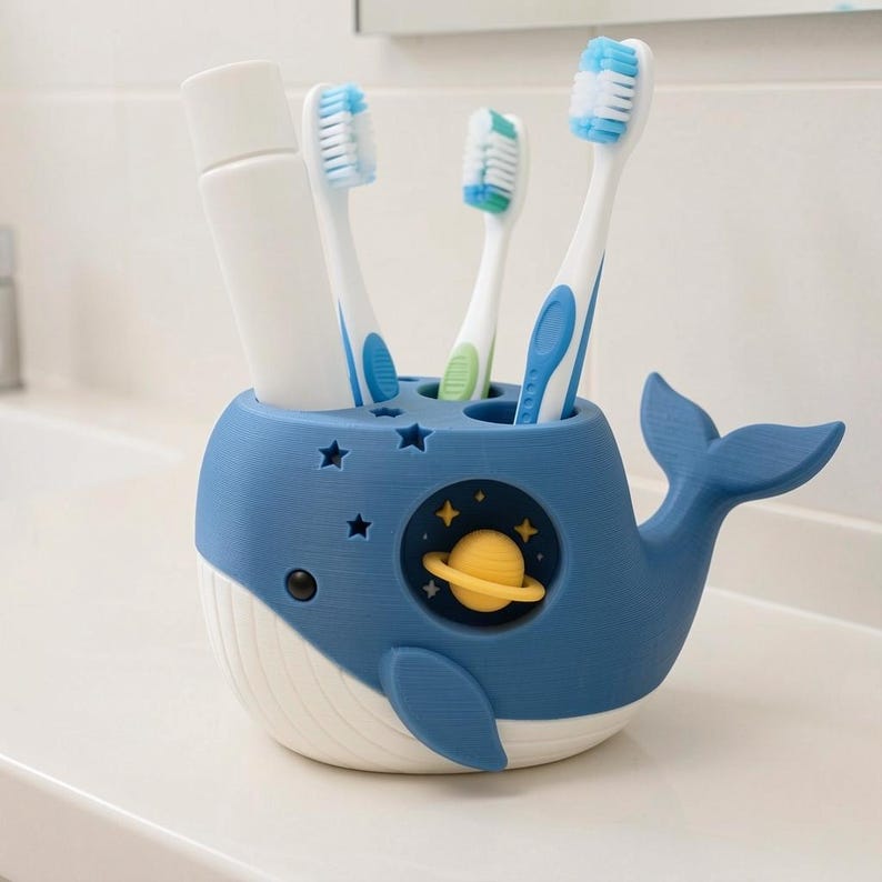 May include: A blue and white whale-shaped toothbrush holder featuring a cutout of a yellow planet with rings and small star-shaped perforations. The holder contains three toothbrushes with blue and white bristles and a white tube of toothpaste.