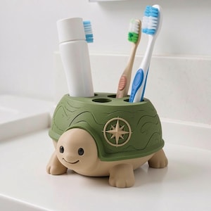 May include: A turtle-shaped toothbrush holder in olive green and beige. The shell features a compass rose emblem and four circular openings. It holds a white tube of toothpaste and two toothbrushes with blue and green bristles, respectively.