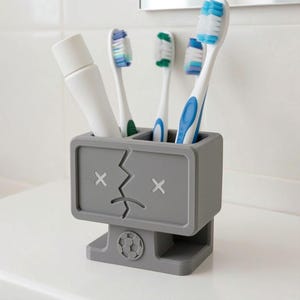 Puede incluir: Un soporte rectangular gris para cepillos de dientes con un rostro estilizado de ojos en forma de X y boca agrietada. La base incluye un emblema circular de bal&oacute;n de f&uacute;tbol. Contiene un tubo de pasta dental blanco y tres cepillos.