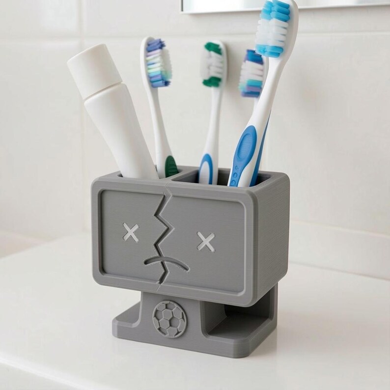 Puede incluir: Un soporte rectangular gris para cepillos de dientes con un rostro estilizado de ojos en forma de X y boca agrietada. La base incluye un emblema circular de bal&oacute;n de f&uacute;tbol. Contiene un tubo de pasta dental blanco y tres cepillos.