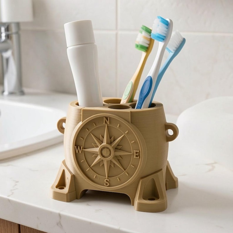 Pode incluir: Um suporte cil&iacute;ndrico para escovas de dentes na cor bege, com um design de rosa dos ventos em relevo na parte frontal, exibindo os pontos cardeais N, E, S e W. Cont&eacute;m um tubo de pasta de dentes branco e tr&ecirc;s escovas com cerdas coloridas.