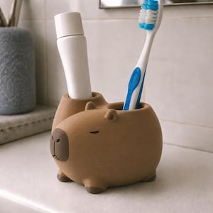 May include: A brown capybara-shaped toothbrush holder with two cylindrical openings. One opening contains a white toothpaste tube, and the other holds a white and blue toothbrush. The holder features a dark brown nose and small ears.