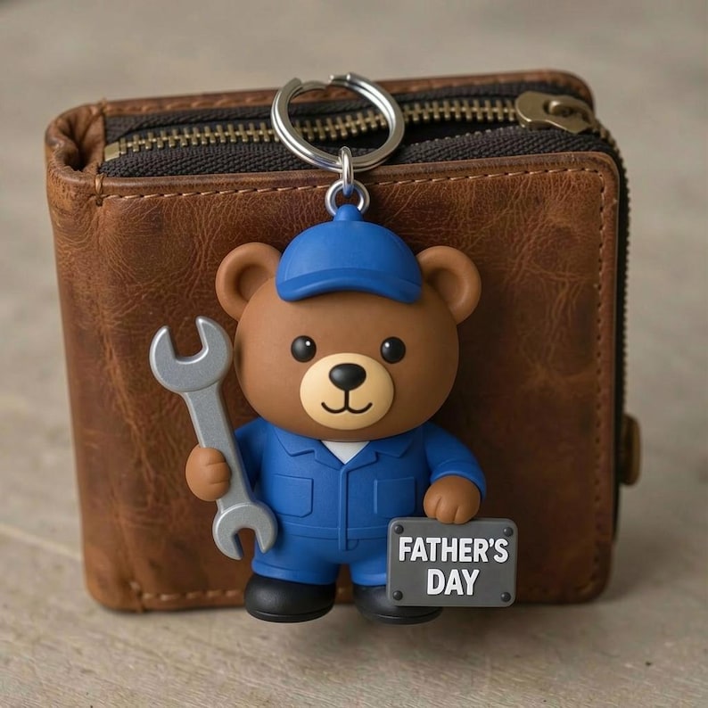 May include: A brown bear keychain wearing a blue cap and blue work uniform, holding a silver wrench in one hand and a gray rectangular sign that reads FATHER'S DAY in the other, positioned in front of a brown leather wallet with a zipper.