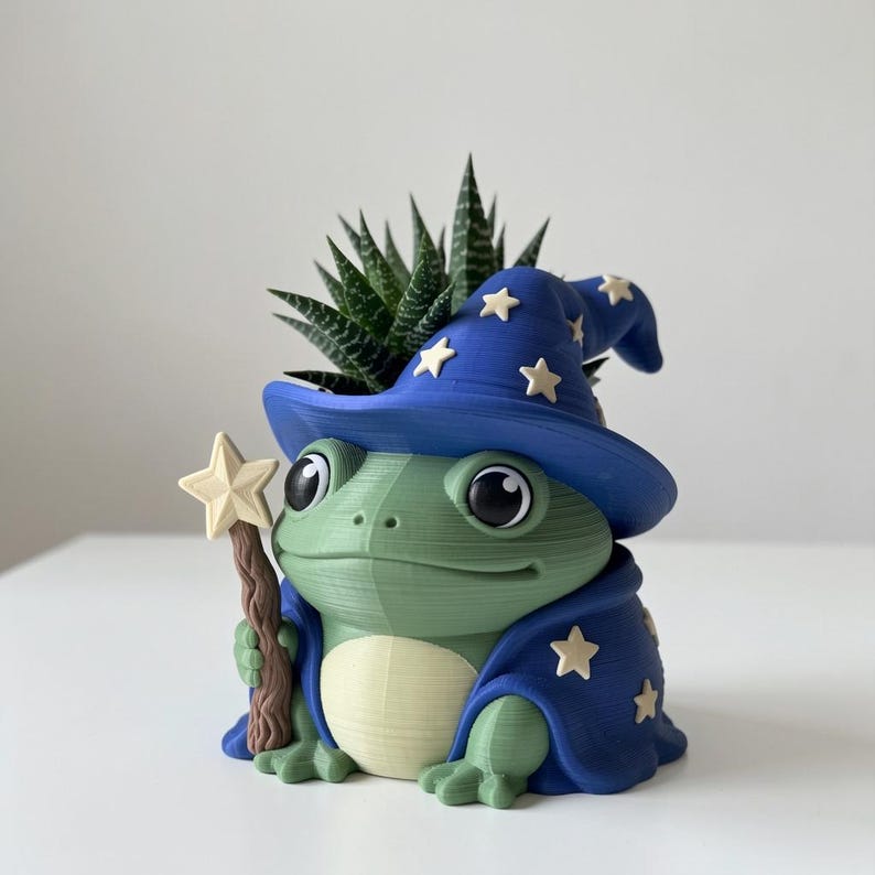May include: A green frog-shaped planter wearing a blue wizard hat and cape decorated with cream-colored stars. The frog holds a brown, textured staff topped with a cream star. A small, spiky green succulent plant grows from the top of the hat.