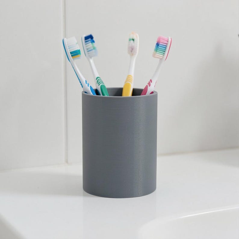 May include: A cylindrical gray toothbrush holder containing four toothbrushes with multicolored bristles. The holder has a smooth, matte finish and a simple, upright design, providing a stable base for storing oral hygiene tools in a bathroom.