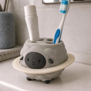 May include: A gray sheep-shaped toothbrush holder with a dark gray face, small ears, and a cream-colored ring encircling its body. The holder features four circular openings on top, currently containing a white tube and a blue and white toothbrush.